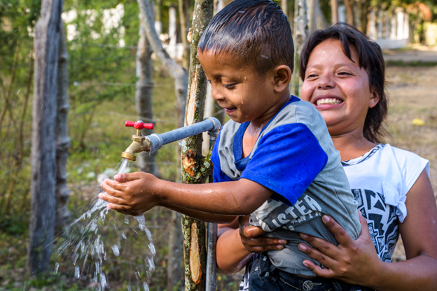 Older child holding younger child up to outdoor faucet.