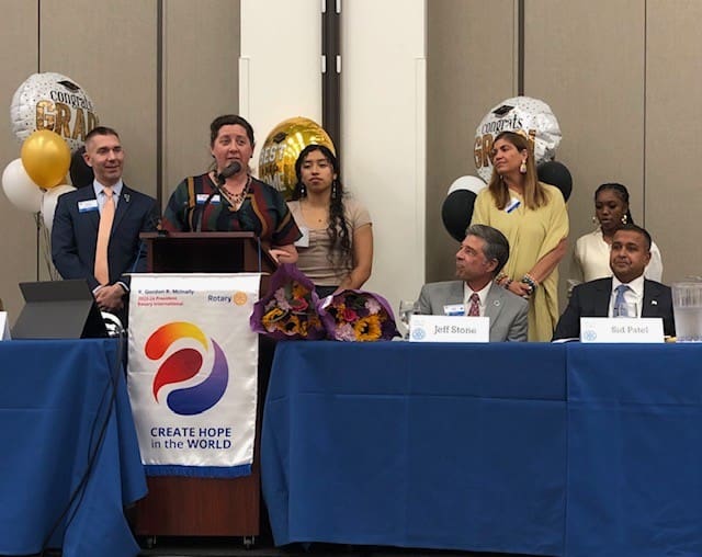 8 people looking at someone speak into a microphone at a stage