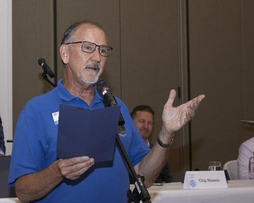 person smiling while speaking into microphone and holding a paper with 1 hand