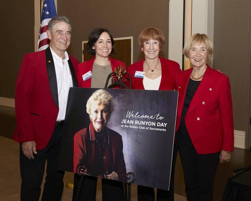 Some men and women of Rotary Sacramento standing together for a photo while wearing red sport coats in front an event poster