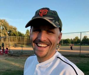 A man smiles at the camera in a Sacramento Rotary club team shirt in front of a soft ball diamond