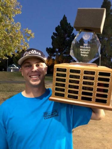 A young man in baseball cap and team Tshirt poses with a softball trophy