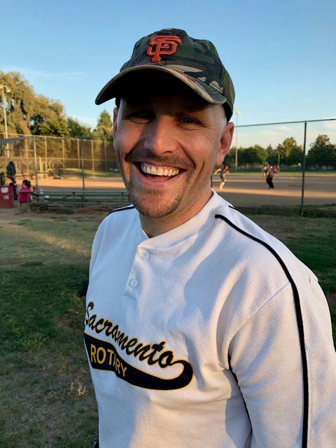A man smiles at the camera in a Sacramento Rotary club team shirt in front of a soft ball diamond