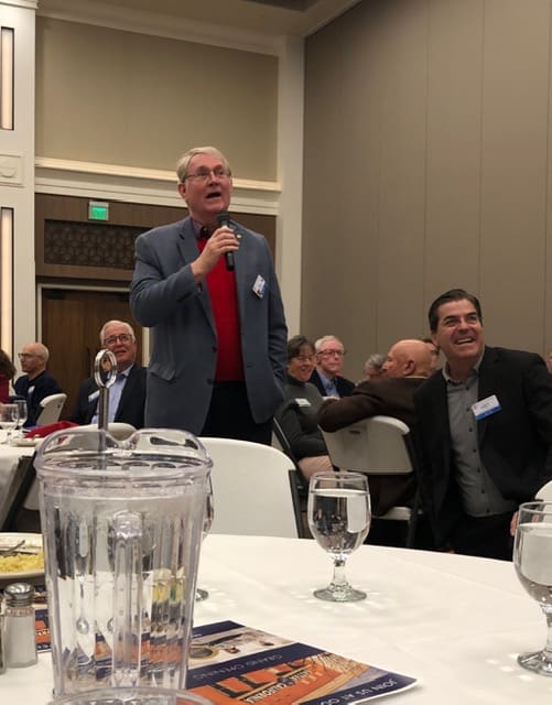 person at a table with a microphone standing up to speak