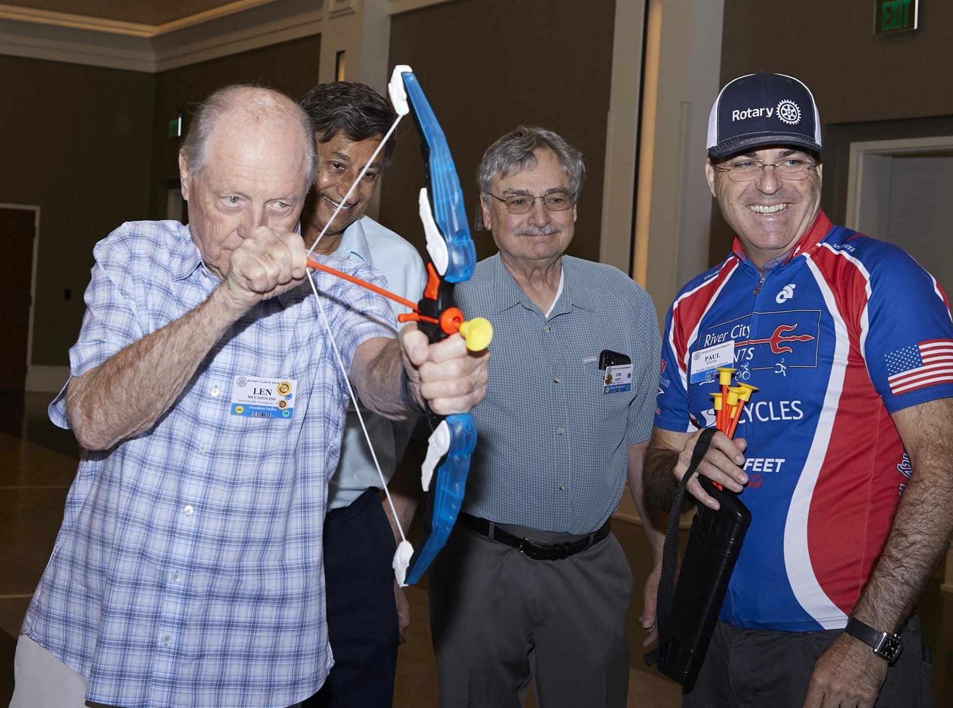 4 people smiling and looking at something off camera while 1 points a toy bow and arrow at something off camera