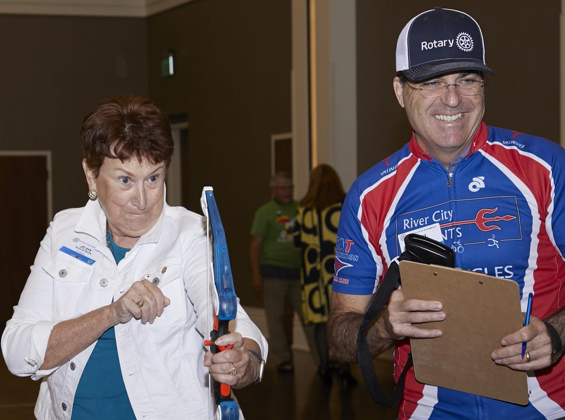 2 people smiling while 1 points a toy bow and arrow at something off camera and the other holds a clipboard