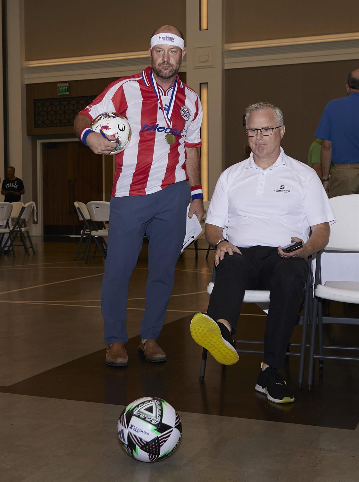 a person standing holding a soccer ball while another people sitting on a chair kicks a soccer ball